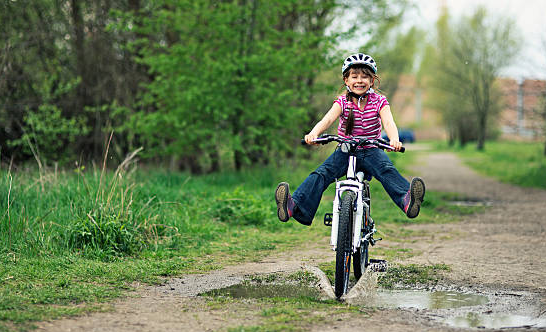 Child riding a bicycle through a small puddle on a dirt path, lifting both legs off the pedals, surrounded by green grass and trees.