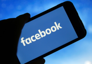 A hand holds a smartphone displaying the Facebook logo on a blue screen, with a blurred blue background behind it.