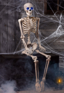 A plastic skeleton with glowing blue eyes sits on a dark wooden chest surrounded by cobwebs, with a small lantern glowing beside it and mist drifting across the floor, creating an eerie Halloween scene.