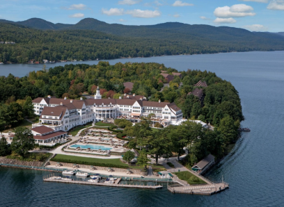  
A large lakeside resort complex on Green Island in Lake George, New York, featuring white buildings with brown roofs, outdoor pools, landscaped grounds, and a long dock extending into the lake. The resort is surrounded by dense green forest and distant mountains under a clear sky.
