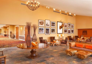 A spacious hotel lobby with rustic décor, featuring a large round wooden table with decorative vases, comfortable seating areas with earth‑tone armchairs and sofas, stone tile flooring, framed photos on the wall, and a mounted deer head creating a cozy lodge‑style atmosphere.