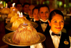   
A row of formally dressed servers carrying dome‑shaped Baked Alaska desserts with browned meringue and small blue flames on top, presenting them in a coordinated procession during a banquet. The warm lighting and flames create a dramatic, celebratory atmosphere.
