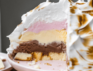 A close-up slice of Baked Alaska showing layers of sponge cake, chocolate, vanilla, and strawberry ice cream, all covered in a thick coating of toasted meringue with golden-brown peaks.