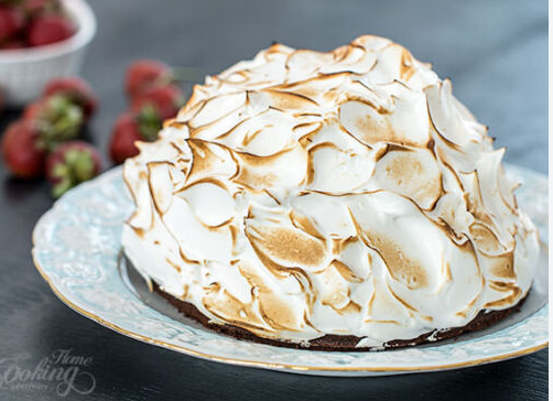 A dome-shaped Baked Alaska covered in toasted meringue with golden-brown peaks, sitting on a decorative plate with fresh strawberries blurred in the background.