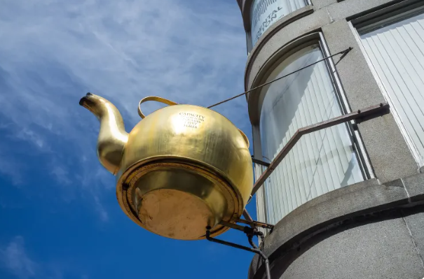 A large golden teapot is mounted on the exterior of a curved stone building, supported by metal brackets. The shiny teapot stands out against a bright blue sky and tall windows, creating a striking landmark on the city street.