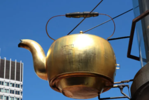   
A large golden teapot hangs outdoors against a blue sky, suspended by metal supports beside a modern building. Text on the side lists its measured capacity in gallons, quarts, pints, and ounces.