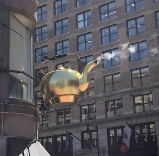 A large golden teapot sculpture hangs from the corner of a downtown Boston building, with real steam rising from its spout. The teapot is mounted high above the sidewalk against a brick façade with rows of rectangular windows.