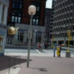   
A large golden teapot hangs from the corner of a downtown building while pedestrians walk below. The street is lined with multi‑story buildings, including one with a “State Street Bank” sign, creating a busy urban scene.