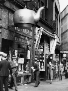   
A vintage street scene shows pedestrians in early‑20th‑century clothing walking past a storefront with a large teapot mounted above the entrance. The shop’s sign reads “Tea Company” and “Home of Best New England Teas,” and surrounding buildings display period advertisements, creating a lively old‑Boston atmosphere.