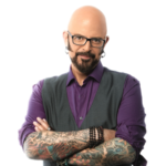 A bald man with a beard and glasses stands against a plain white background. He wears a purple shirt with rolled‑up sleeves, a gray vest, and multiple bracelets. His tattooed arms are crossed as he looks directly at the camera with a confident expression.