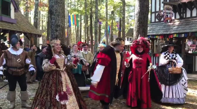 Group of people dressed in ornate Renaissance-style costumes standing outdoors among tall trees. They wear velvet and brocade garments with crowns, hats, and capes. Timber-framed buildings decorated with colorful banners are visible in the background at a renaissance fair.”