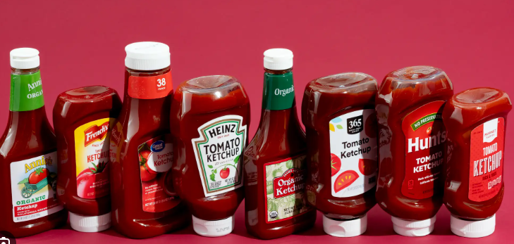   
Eight different ketchup bottles from various brands arranged side by side against a solid red background, showing the variety of packaging styles and labels.
