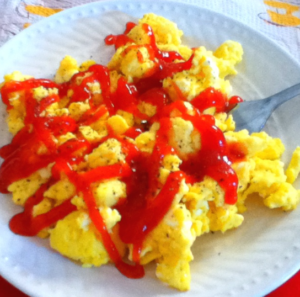   
A plate of fluffy scrambled eggs topped with a zigzag drizzle of ketchup, with a fork resting beside the eggs.