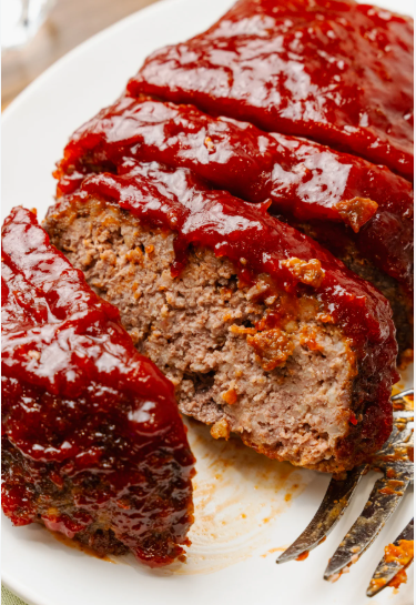 A sliced meatloaf sits on a white plate, topped with a thick, glossy red glaze. The inside looks moist and finely textured, with small bits of seasoning visible. A fork rests near the edge of the plate, suggesting it’s ready to be served.