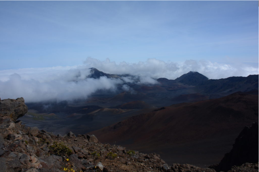   
“Rugged volcanic crater landscape with rocky terrain in the foreground and tall peaks rising above drifting clouds under a bright blue sky.”