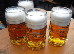 Four glass beer steins filled with golden Märzen beer topped with thick foam, sitting on a wooden outdoor table at a festive beer garden.