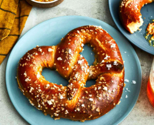  A large golden‑brown soft pretzel sprinkled with coarse salt on a blue plate, with another pretzel, mustard dip, and a napkin blurred in the background.