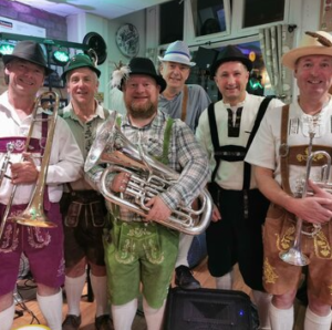 Six musicians dressed in traditional Bavarian clothing holding brass instruments and posing indoors at a festive Oktoberfest-style event.