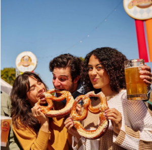 Three people outdoors enjoying large salted pretzels and beer at a sunny festival. Colorful banners and signs in the background suggest an Oktoberfest celebration, with bright light and a cheerful atmosphere