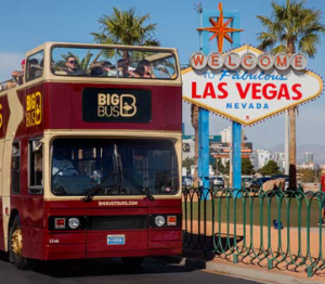   
Big Bus double‑decker tour bus passing the Welcome to Fabulous Las Vegas sign on a sunny day, with palm trees and blue sky in the background.