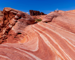 Layered red and pink sandstone with smooth wave‑like striations set against dark rock formations and a bright blue desert sky.