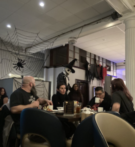 Diners seated at a Halloween‑decorated restaurant table surrounded by oversized spider webs, hanging spiders, and life‑size skeleton figures, including one dressed like Jack Skellington in a witch hat.