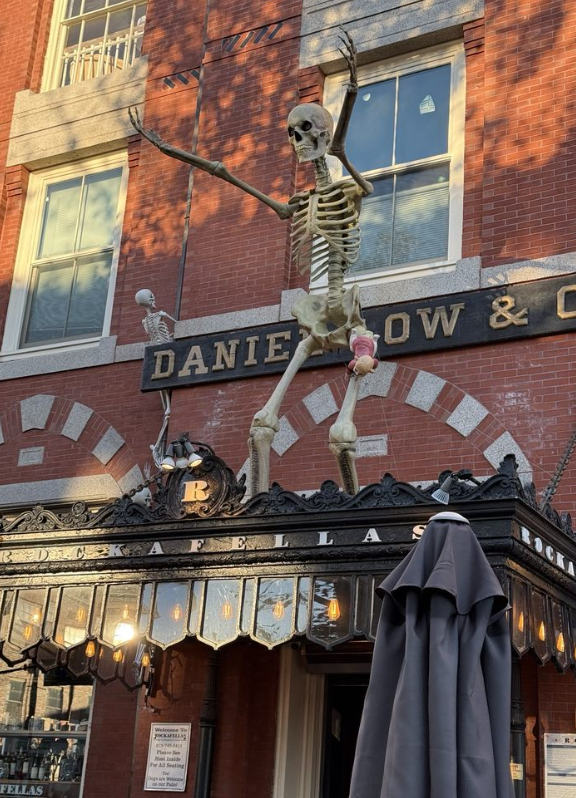 A historic brick building in Salem with a large skeleton decoration mounted above the entrance, signage reading “Daniel Low & Co.” and “Rockafellas,” and additional Halloween skeletons near the upper windows.