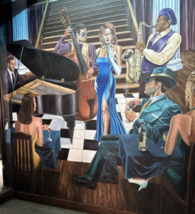 A vibrant mural depicting a jazz club scene, featuring a woman in a flowing blue dress singing on a checkered floor, surrounded by musicians playing piano, bass, and saxophone, with two patrons seated at a table in the foreground.