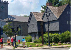 A group of visitors standing near the historic Witch House in Salem, a dark wooden 17th‑century structure with steep gables, set beside a large Gothic stone building with arched windows and battlements
