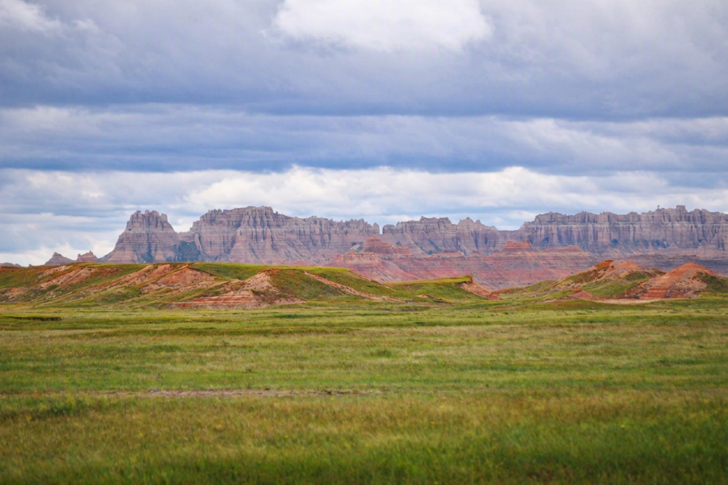 Expansive badlands landscape with layered rock formations, rugged cliffs, and grassy rolling hills under a partly cloudy sky.