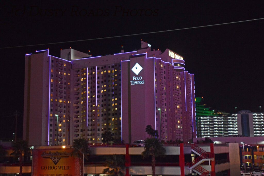 Nighttime view of the Polo Towers high‑rise on the Las Vegas Strip, illuminated in purple and white lights with palm trees and the Harley‑Davidson Café sign in the foreground.