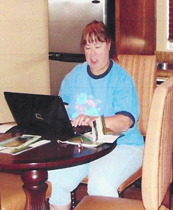 Person sitting at a round wooden table working on a laptop, with papers and an open book nearby in a casual indoor setting.
