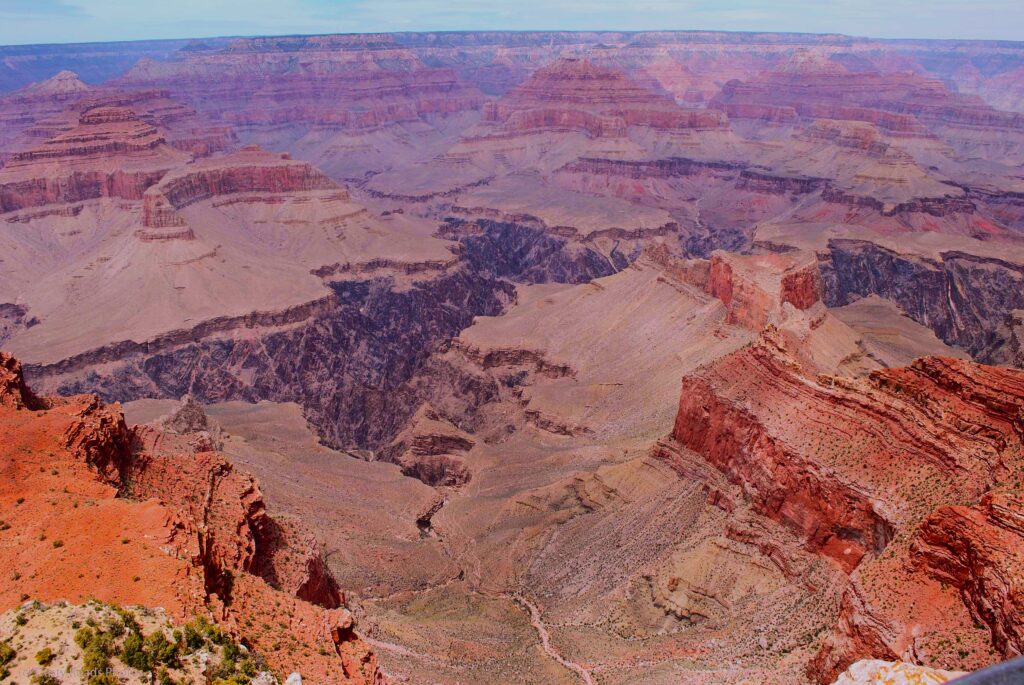 Expansive view of the Grand Canyon with layered red and orange cliffs, deep valleys, and rugged rock formations stretching toward the horizon under a partly cloudy sky.