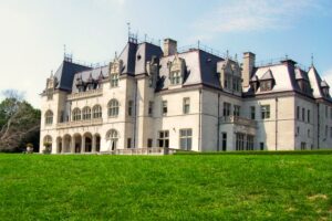 A grand seaside mansion rises above a wide green lawn, viewed from the Cliff Walk. The light‑colored stone building features steep gabled roofs, ornate chimneys, and arched windows under a bright, lightly clouded sky.