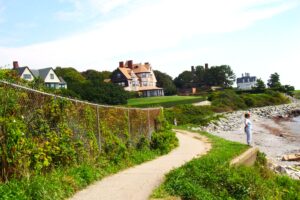 A coastal walking path curves along the shoreline, bordered by greenery and a wire fence. Large seaside homes sit on the hill above, and a person stands near the water’s edge looking out toward the ocean under a bright, peaceful sky.