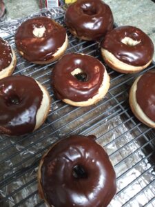 Seven chocolate‑glazed donuts rest on a metal cooling rack set over aluminum foil, their glossy tops contrasting with the golden‑brown dough beneath.