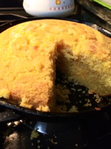   Golden cornbread baked in a black cast‑iron skillet, with a large wedge cut out to show its moist, crumbly interior, sitting on a stovetop.