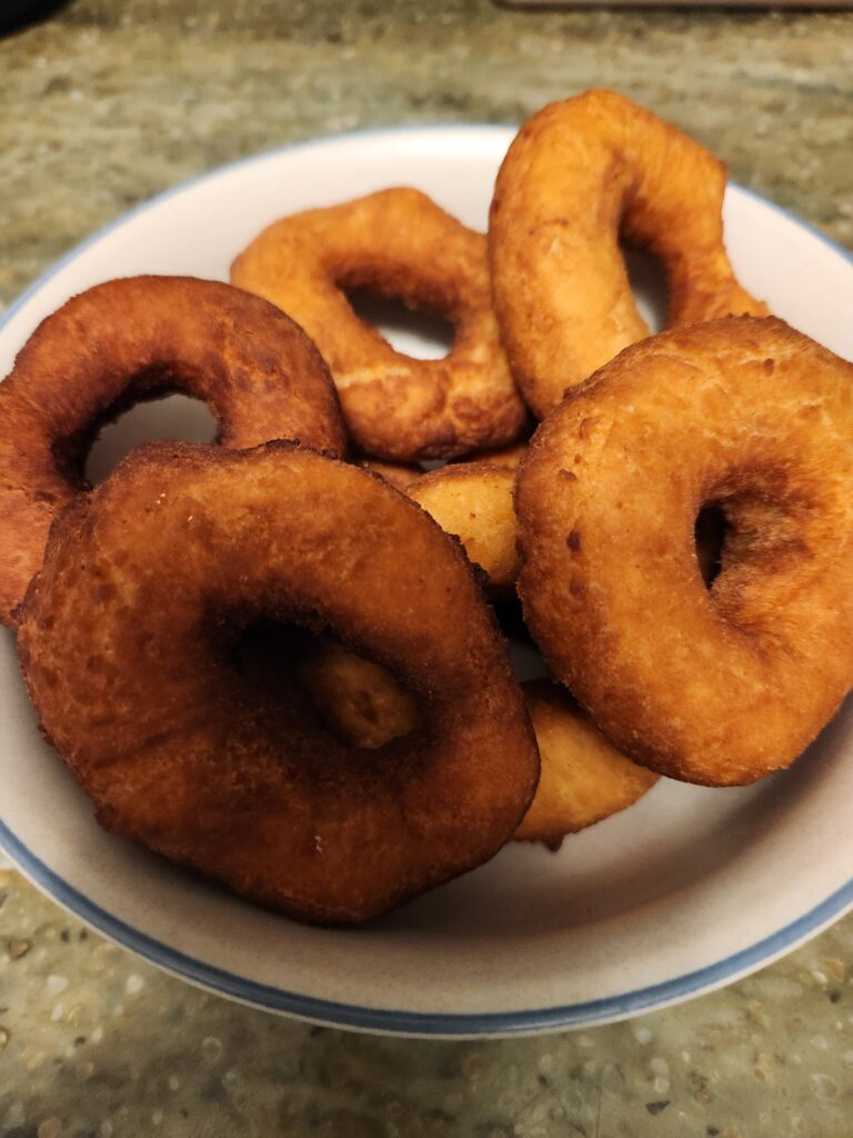 A white plate with a blue rim holds several homemade, golden‑brown fried donuts with slightly irregular shapes, sitting on a kitchen countertop.