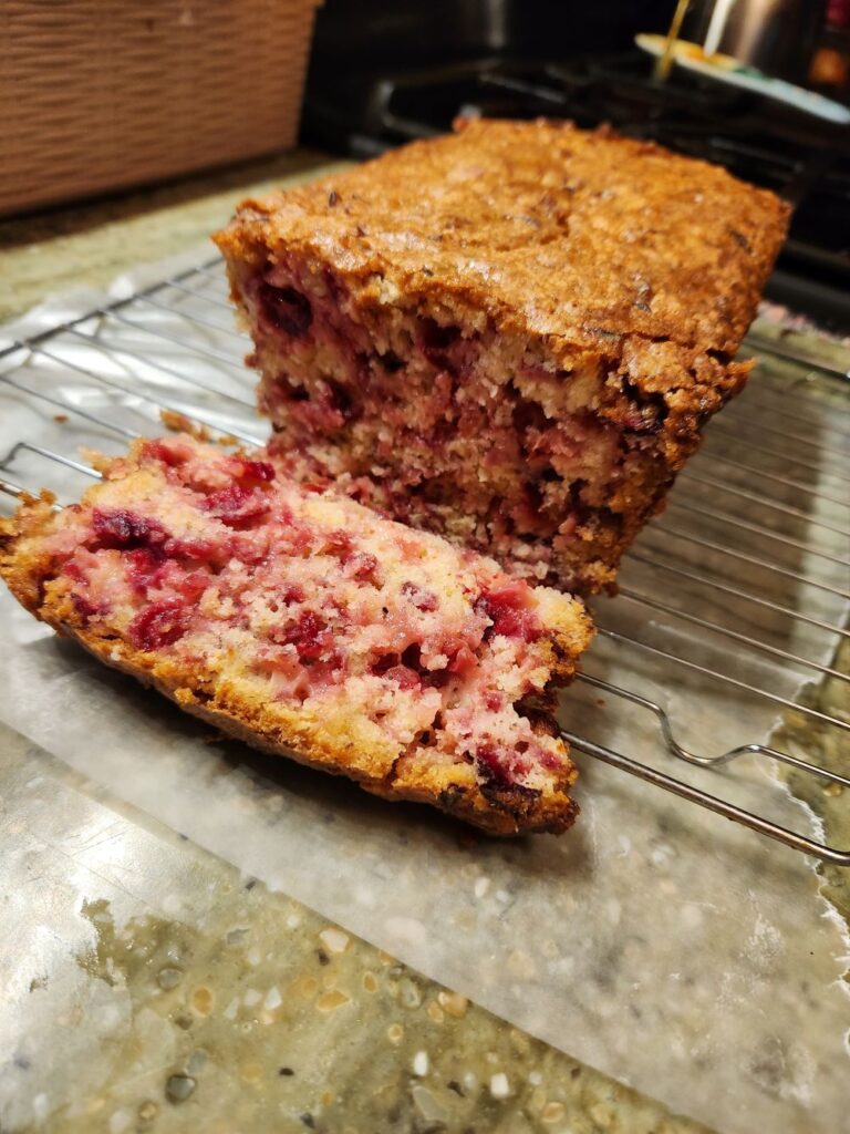 “Homemade cranberry bread loaf on a cooling rack with one slice cut, showing a golden crust and moist interior with bright red berries.”
