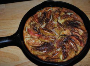 A classic Bickford‑style apple pancake baked in a black cast‑iron skillet, with thin apple slices arranged in a circular pattern over a golden, custardy base and lightly dusted with cinnamon and sugar.