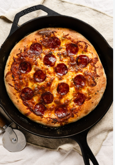 A freshly baked pepperoni pizza in a black cast‑iron skillet, with a golden crust and melted cheese, sitting on a light cloth beside a metal pizza cutter