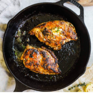   Two golden, oven‑roasted chicken breasts resting in a black cast‑iron skillet, lightly browned with melted butter and herbs, set on a cloth-covered countertop.