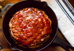 A freshly baked meatloaf in a black cast‑iron skillet, topped with a glossy red ketchup glaze, resting on a wooden surface beside a striped kitchen towel.