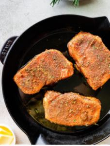   Three seasoned pork chops arranged in a black cast‑iron skillet, coated with a reddish-brown spice rub and ready to cook, with a sprig of rosemary nearby on a light surface.
