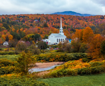  A white steepled church sits in a small Vermont town surrounded by vibrant autumn foliage, with red, orange, and yellow trees, a grassy foreground, a winding stream, and misty mountains rising in the background.