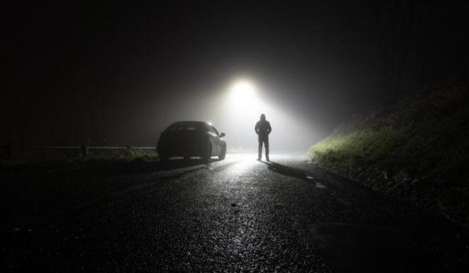  A lone figure stands in the middle of a dark, foggy road at night, illuminated by a harsh overhead light while a parked car sits nearby, its headlights reflecting off the wet pavement to create an eerie, suspenseful atmosphere.
