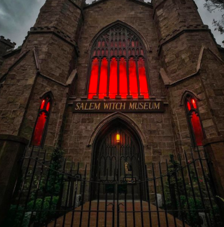 Front entrance of the Salem Witch Museum, a dark stone Gothic building with tall arched windows glowing red from interior lighting. The museum’s sign hangs above the doorway, and an iron gate with surrounding greenery frames the scene, creating an eerie, dramatic atmosphere.