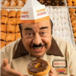 A smiling Dunkin’ Donuts employee in a beige uniform and paper hat holds up a chocolate‑glazed donut while standing in front of trays filled with assorted donuts.