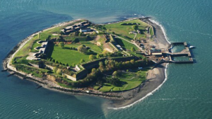   
Aerial view of a small island surrounded by water, featuring a large historic stone fort with bastioned walls, grassy interior grounds, and several buildings arranged within the complex. A long pier extends from one side of the island, highlighting its isolated coastal setting.