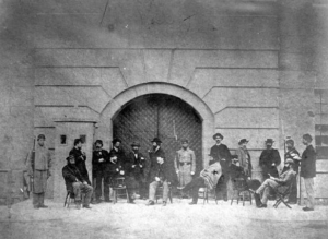   
Black‑and‑white historical photograph showing a group of 19th‑century soldiers and civilians gathered outside a large arched doorway of a stone fort. Some sit on chairs while others stand around them, dressed in period military uniforms and civilian clothing, creating an informal scene of life at the fort.